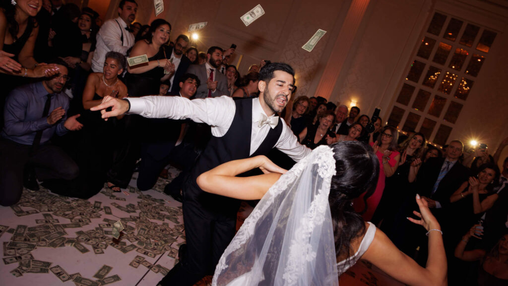 Greek-American groom and bride performing a traditional money dance surrounded by cheering wedding guests